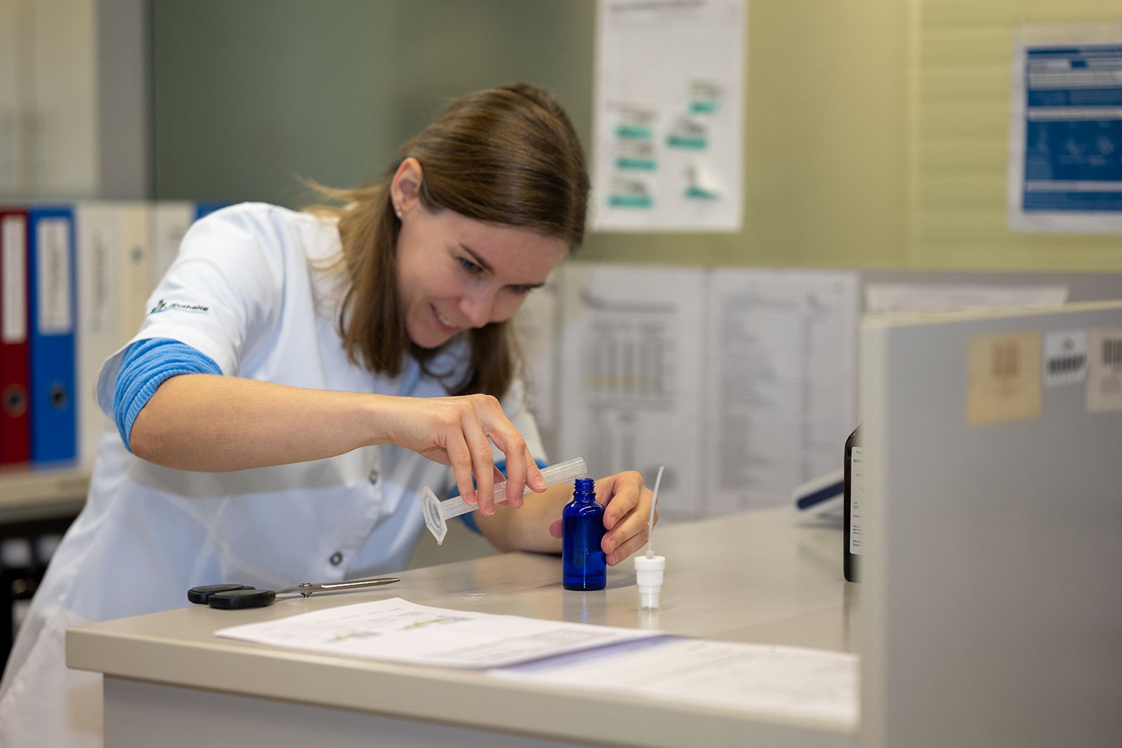 Eine Frau im Laborkittel füllt an einem Tresen eine kleine blaue Flasche mit einer Spritze.
