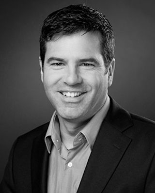 Auf einem Schwarzweißfoto lächelt ein Mann in Anzug und Hemd.