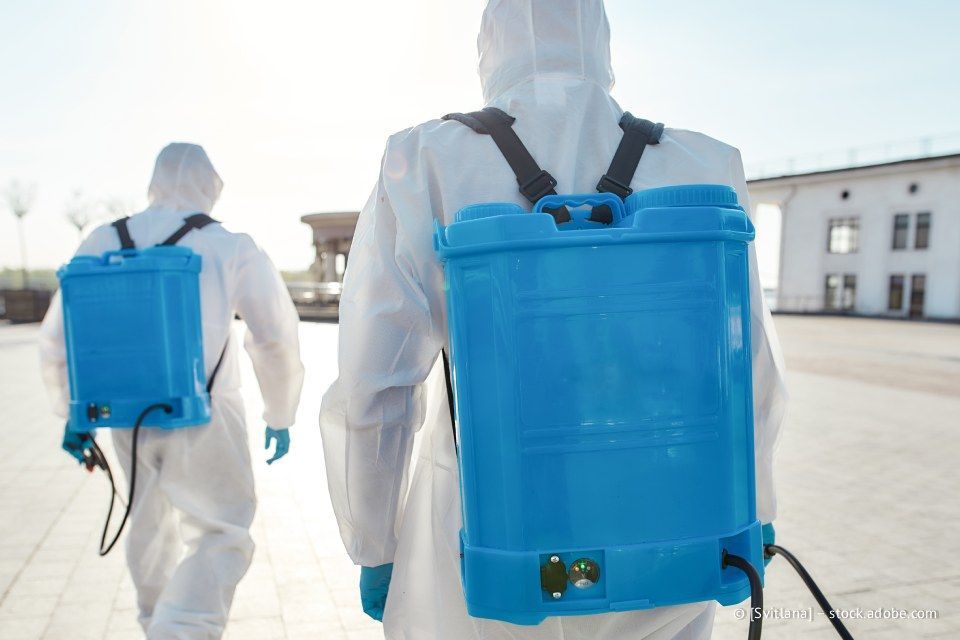 Zwei Personen in weißen Schutzanzügen mit blauen Sprühtanks gehen auf einem gepflasterten Platz.