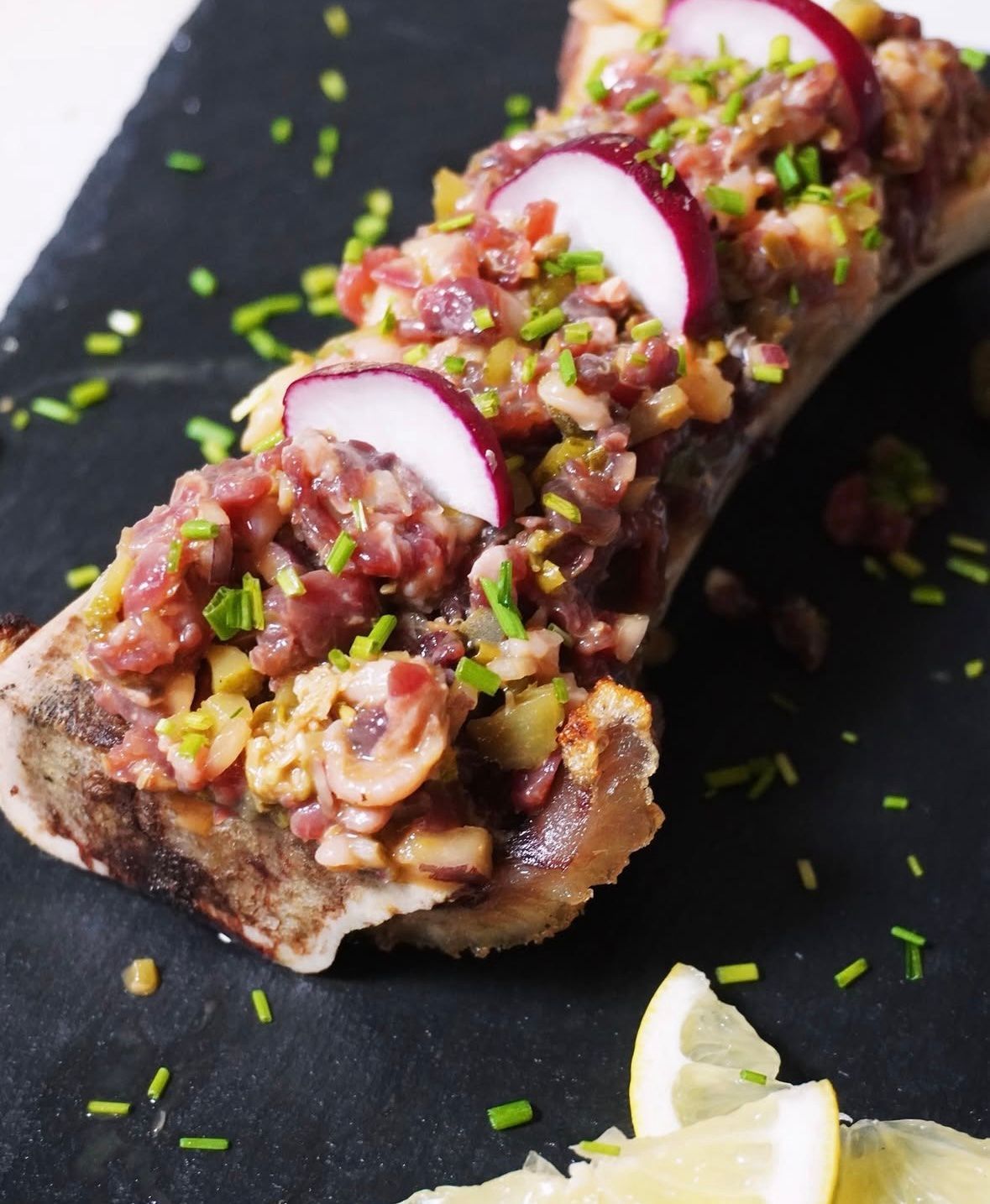 Tarta gourmet cubierta con atún en cubos, rodajas de rábano y cebollino en un plato oscuro con rodajas de limón.