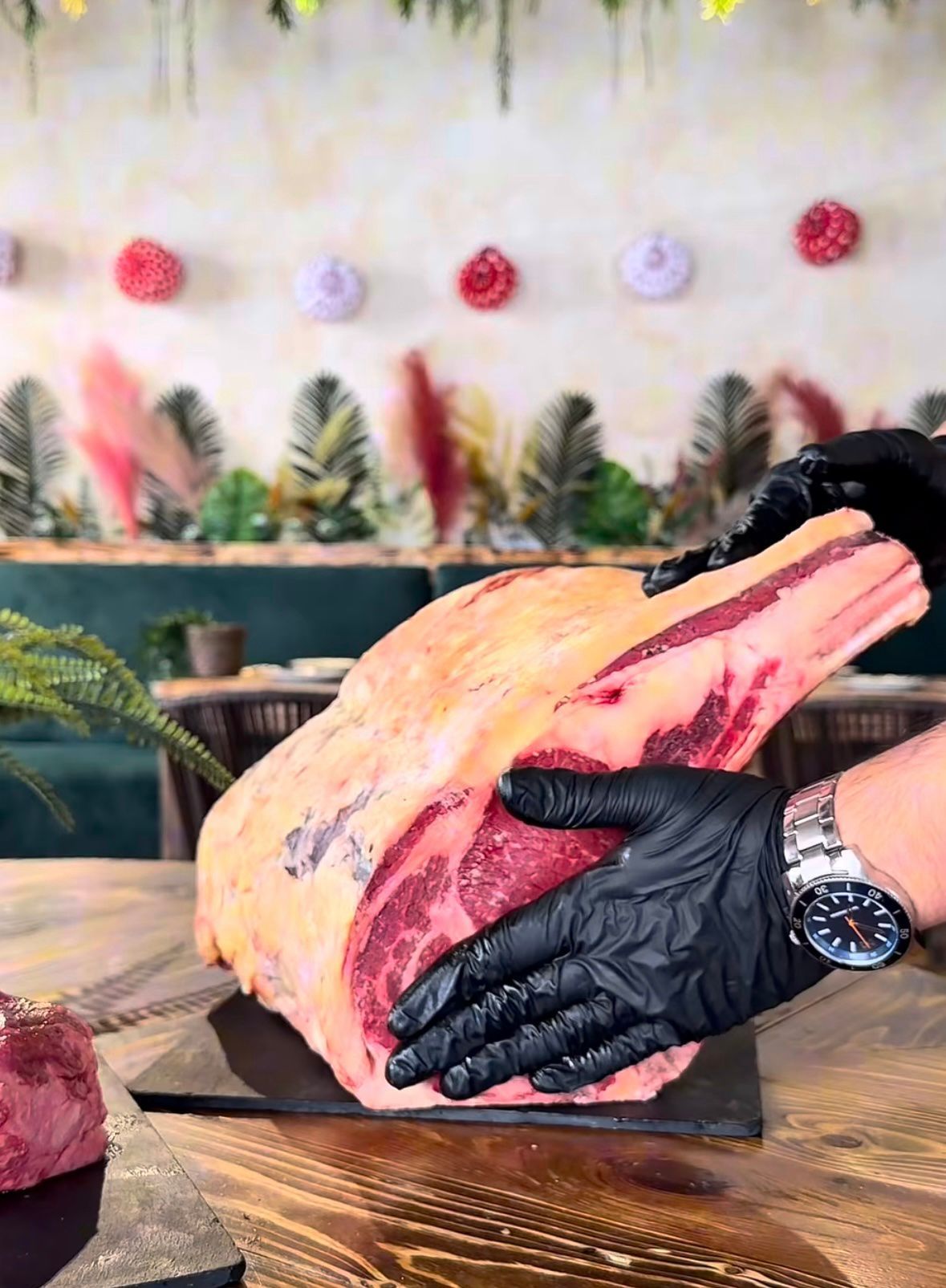 Unas manos enguantadas de negro sostienen un gran trozo de carne cruda veteada sobre una mesa de madera
