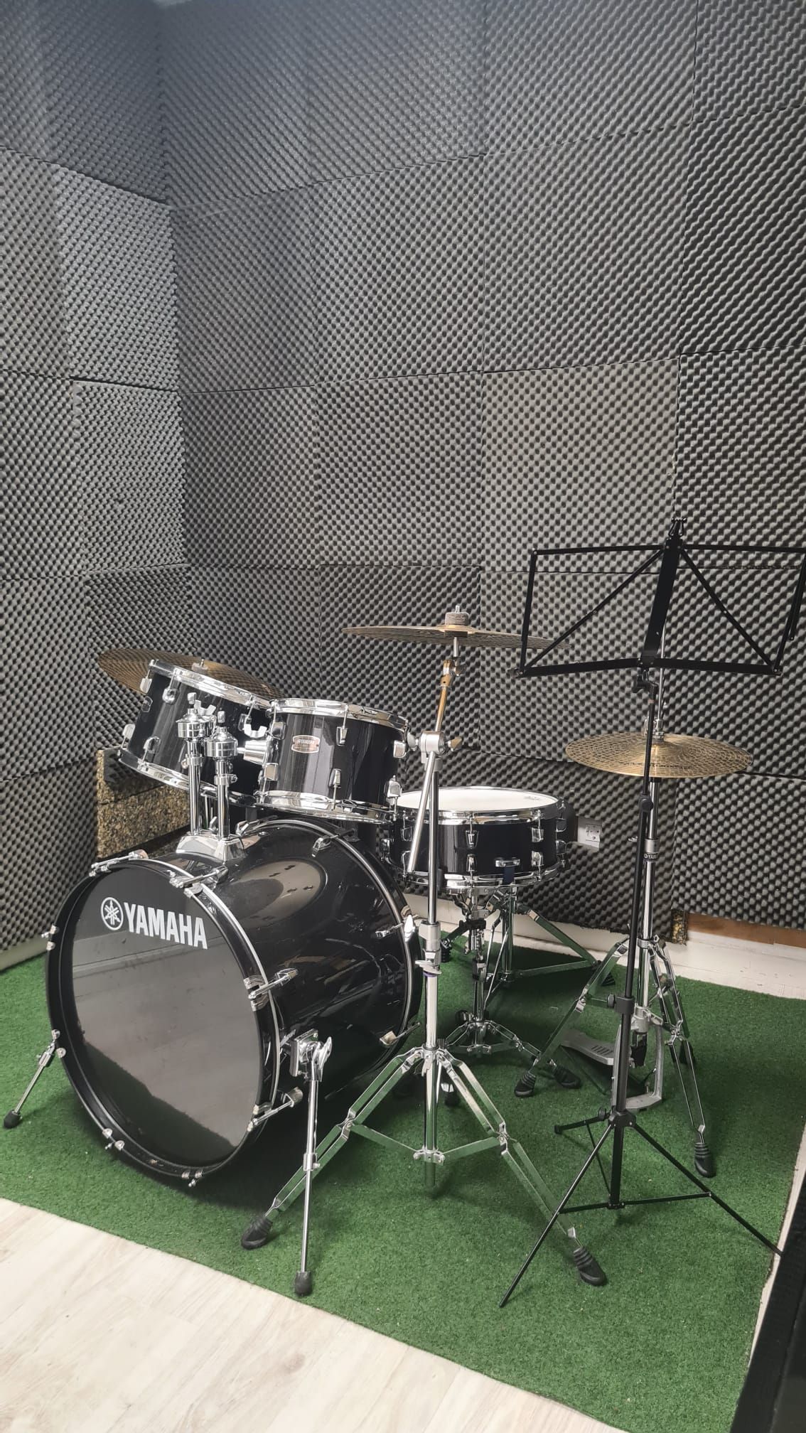 Una batería sobre una alfombra verde en una sala de ensayo musical con paredes de espuma acústica negra y un atril.
