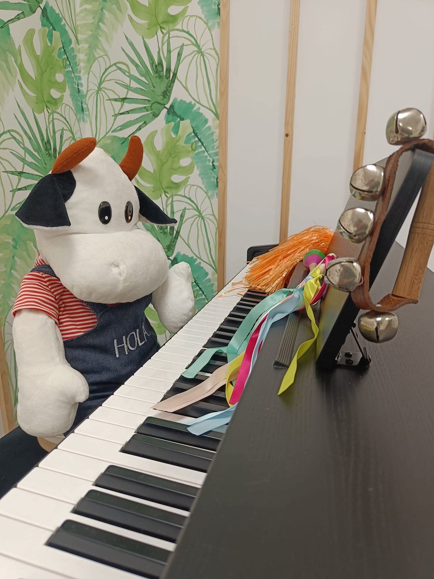 Una vaca de peluche junto a un teclado de piano con cintas de colores sobre una mesa negra, cerca de una figurita de metal.