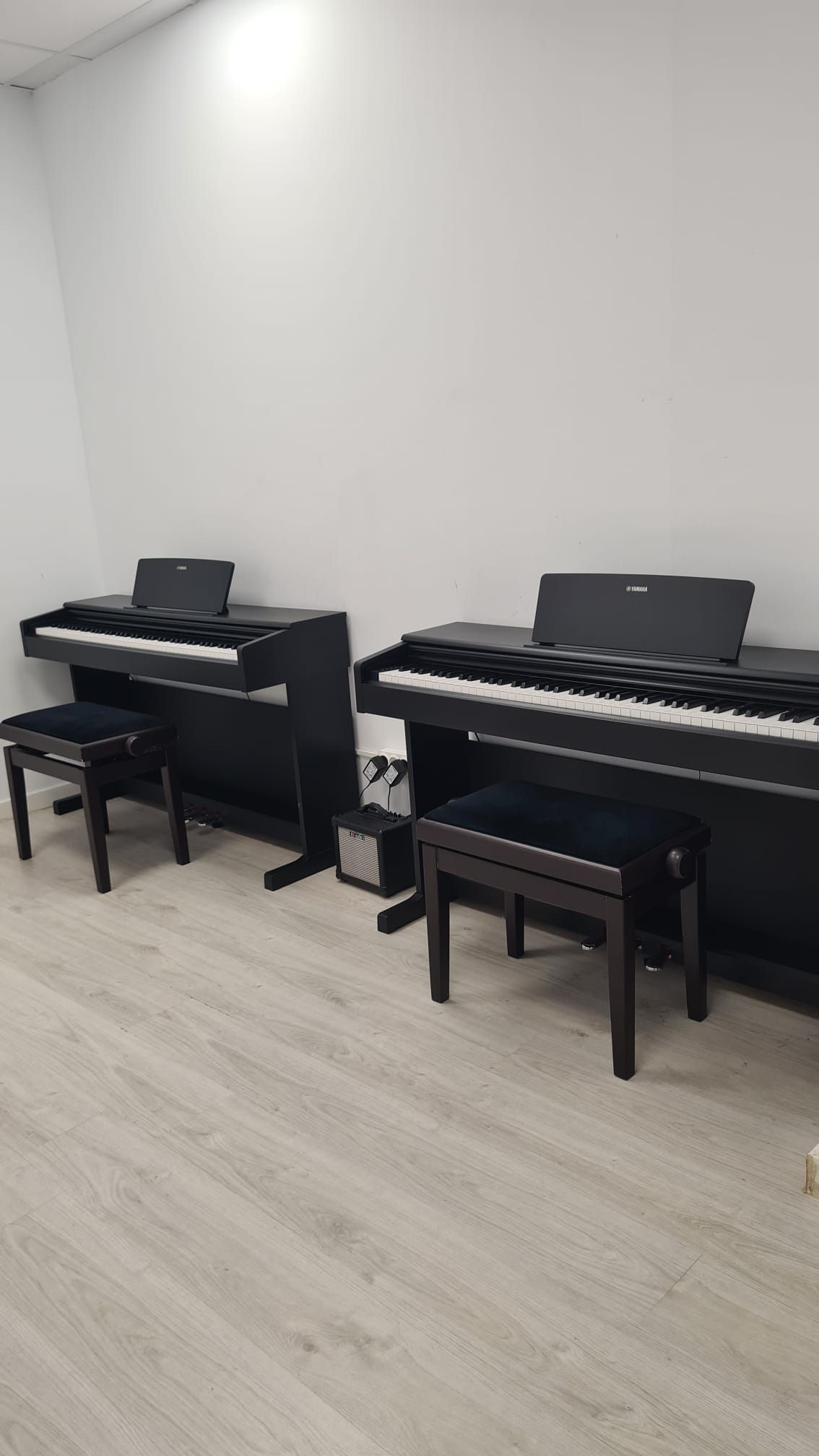 Dos pianos digitales negros con banquetas contra una pared blanca lisa en una habitación luminosa.