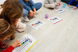 Los niños se sientan en sillas de colores, con las manos levantadas, en un aula donde hay un profesor sentado al frente.