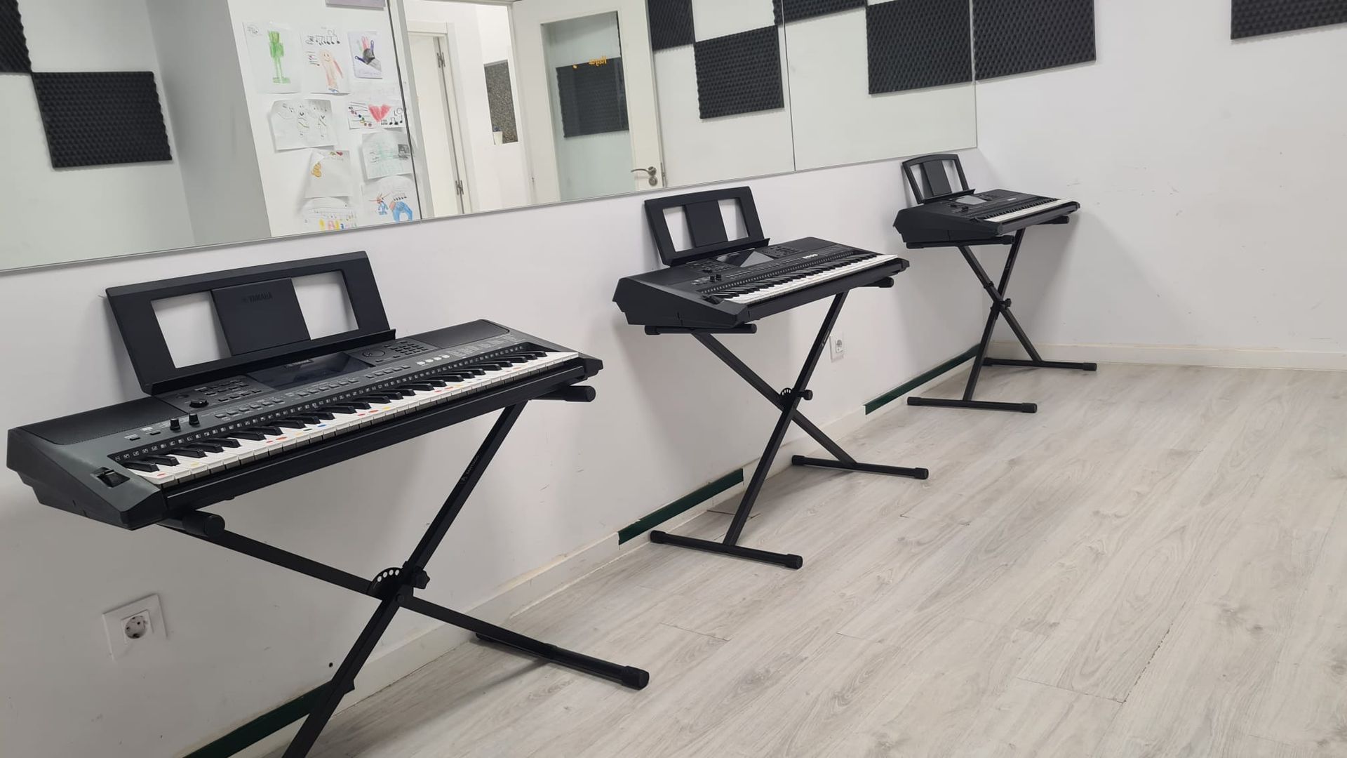 Tres teclados electrónicos sobre soportes en forma de X en un aula de música luminosa.