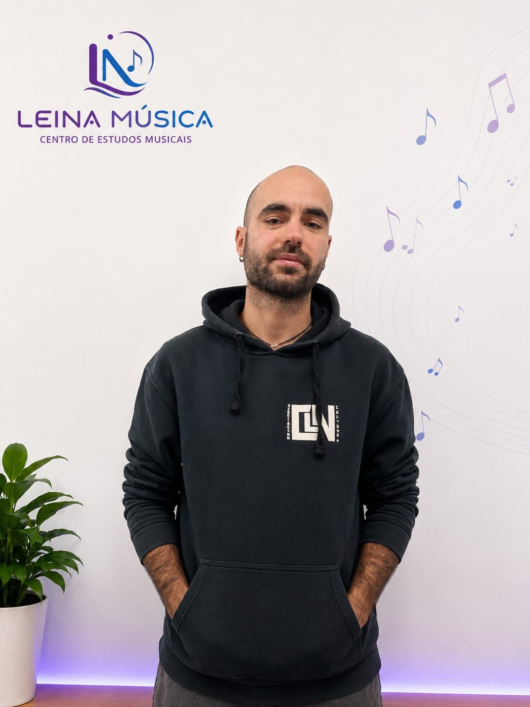 Hombre de pie con una sudadera negra en un estudio de música con el logo de LEINA MÚSICA en la pared.
