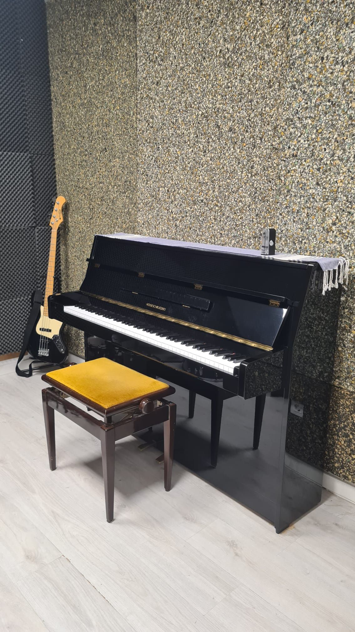 Un piano vertical negro con banqueta y una guitarra en una habitación pequeña con papel tapiz estampado en las paredes.