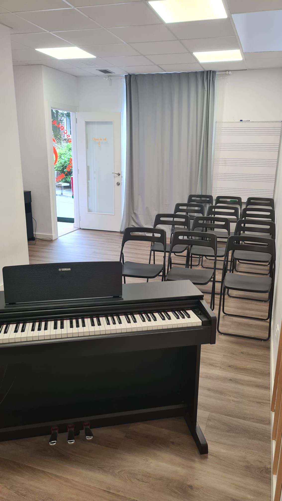 Un piano en primer plano de una pequeña sala de música con sillas apiladas, paredes blancas