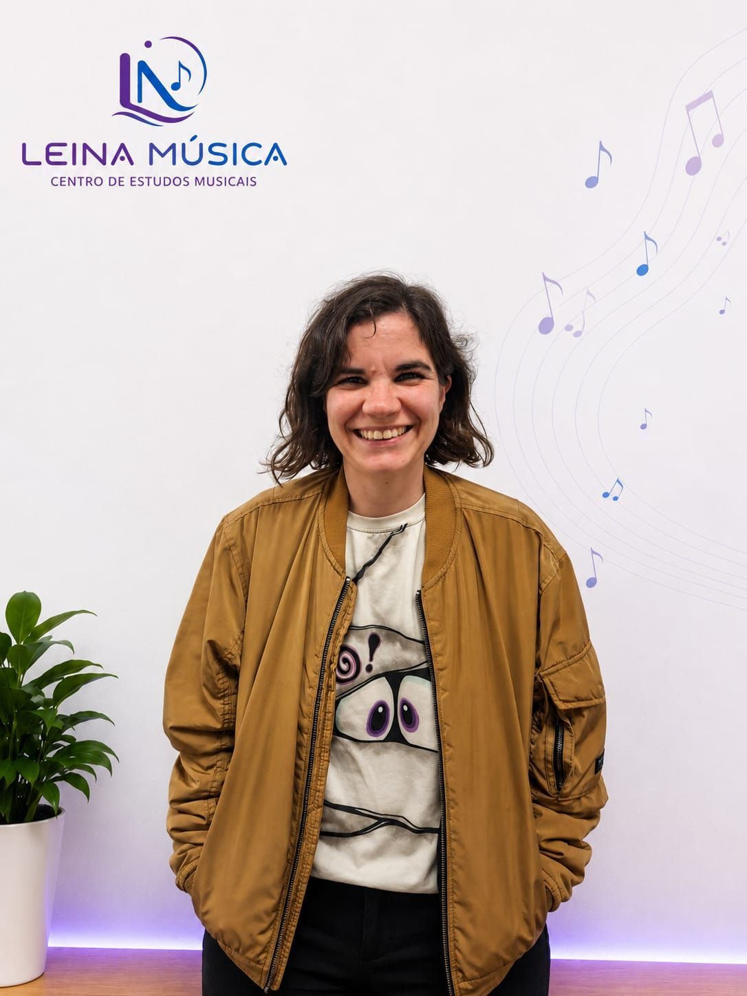 Una mujer sonriente con una chaqueta color canela posa frente a una pared con el logotipo de Leina Música.