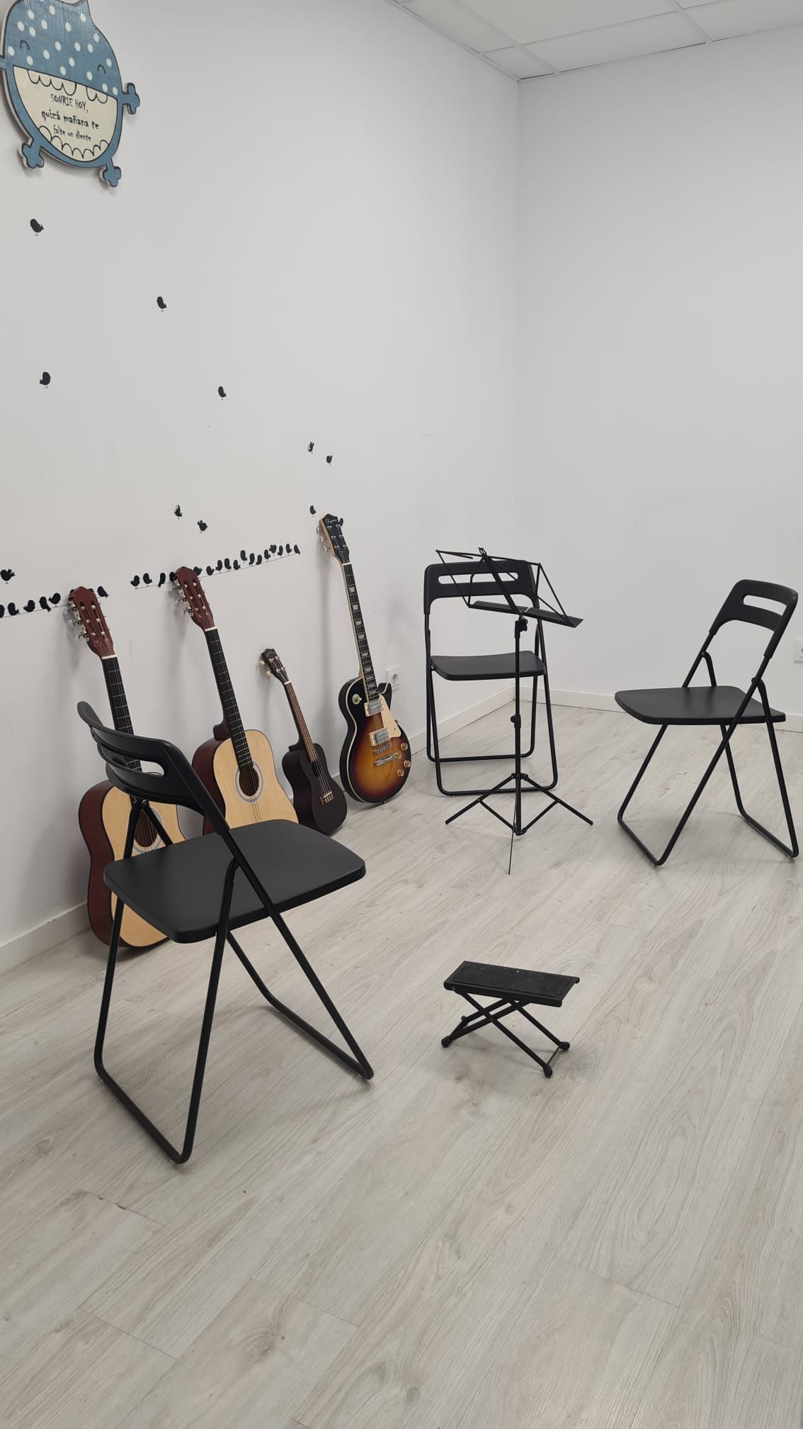 Pequeña sala de ensayo con varias guitarras, una silla plegable, un atril y un taburete sobre un suelo de madera clara.