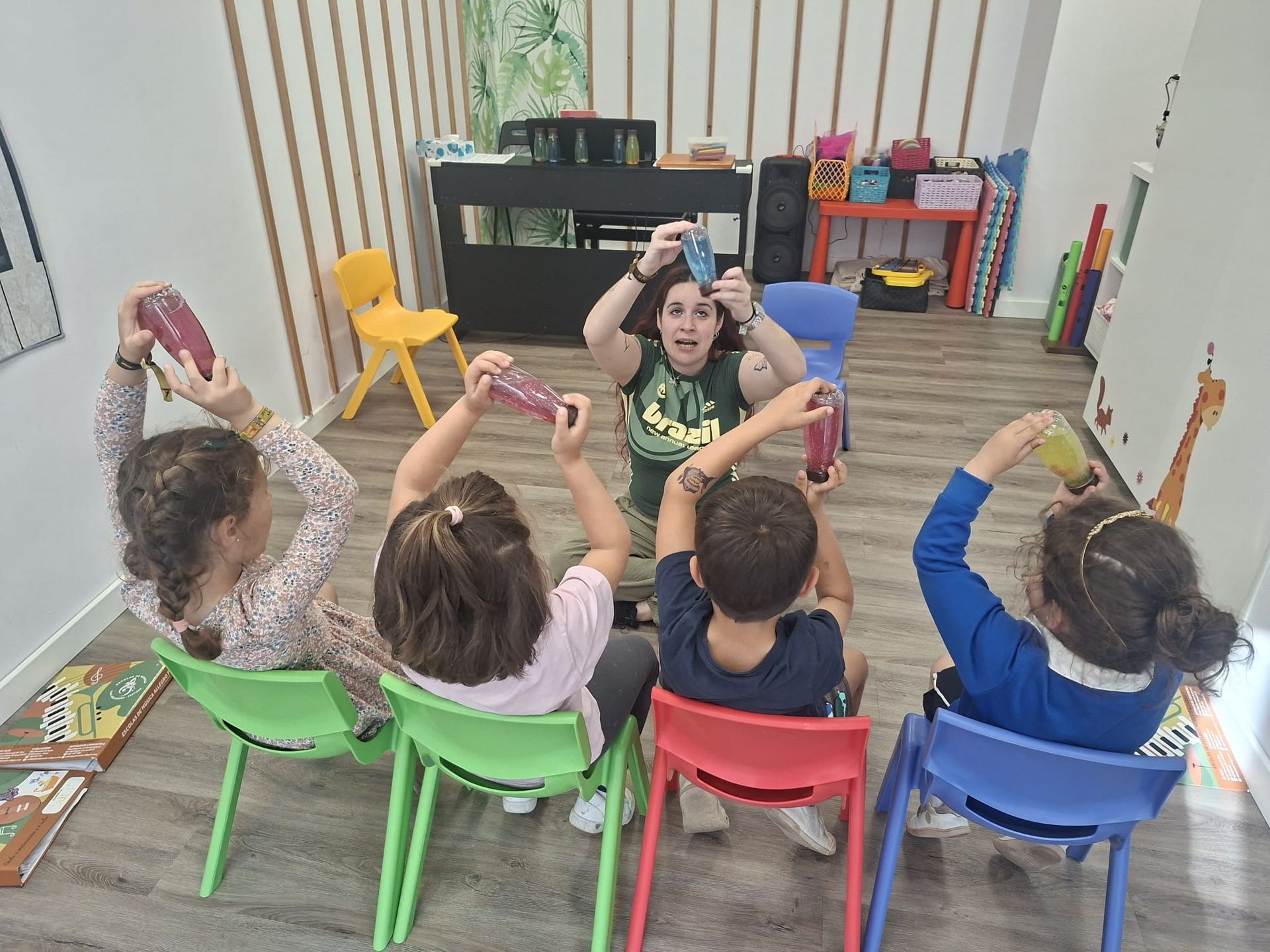 Niños sentados en sillas coloridas, levantando las manos hacia un maestro en una actividad en el aula.