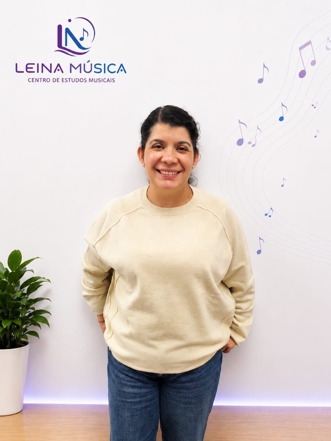Mujer sonriente con jersey beige y vaqueros junto a una pared con el logo de Leina Música, con plantas y notas musicales.