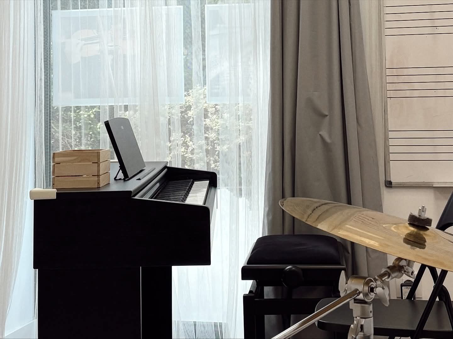 Un piano vertical negro junto a una batería en una habitación luminosa con cortinas transparentes y visillos beige.