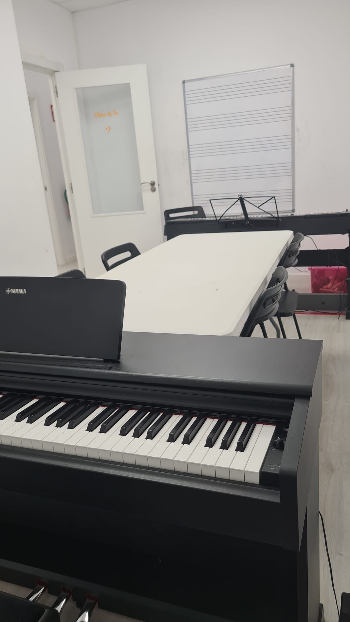 Pequeña sala de música con piano digital, ordenador portátil, mesa plegable y pizarra blanca.