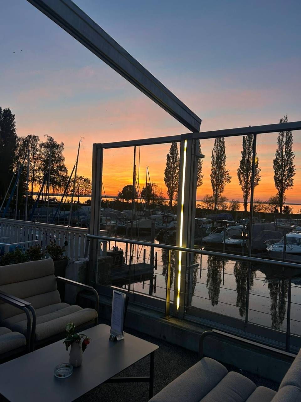 Sonnenuntergang über einer Terrasse am Wasser mit Lounge-Sitzgelegenheiten, Glasgeländer und Spiegelungen im Wasser
