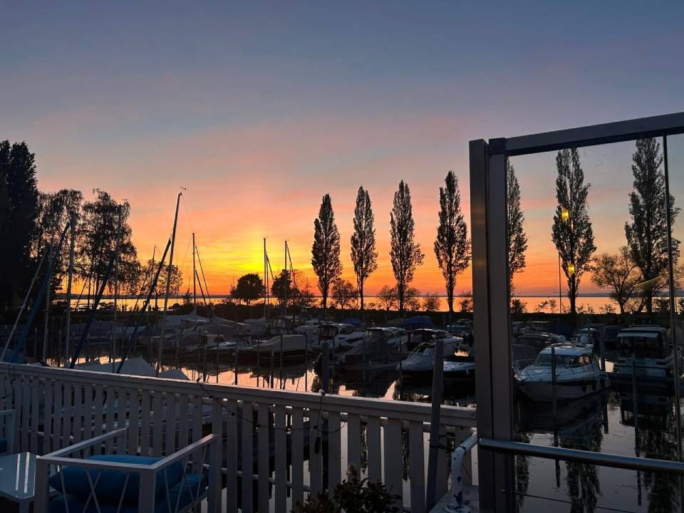 Sonnenuntergang über einem Jachthafen mit silhouettenhaften Bäumen, Booten und einem weissen Geländer im Vordergrund