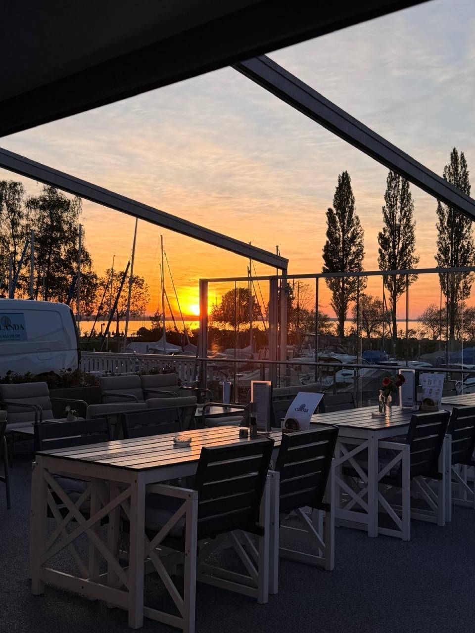 Terrasse eines Restaurants im Freien bei Sonnenuntergang, mit Tischen, Stühlen und sich gegen den orangefarbenen Himmel abzeichnenden Baumsilhouetten.
