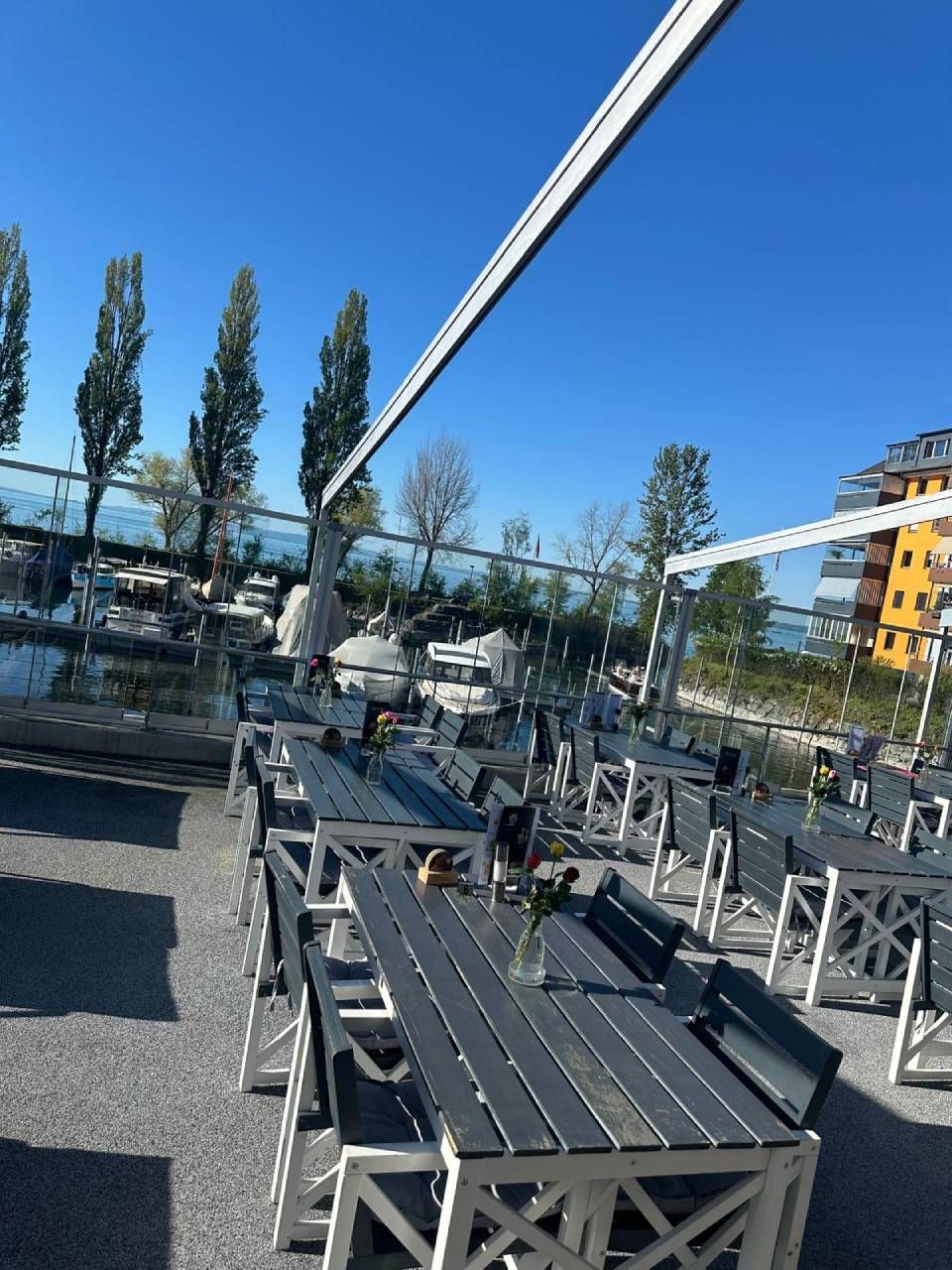Sitzgelegenheiten auf einer Terrasse mit Metalltischen und -stühlen unter blauem Himmel, im Hintergrund ist eine Person zu sehen.