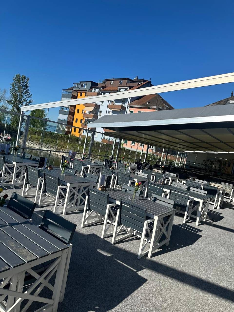 Aussenterrasse eines Cafés mit grauen Tischen und Stühlen unter strahlend blauem Himmel, in der Nähe moderner Gebäude.