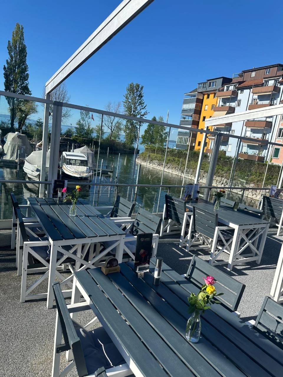 Aussenterrasse eines Cafés mit blauen Tischen, weissen Stühlen, Glasgeländer und Wohnhäusern im Hintergrund