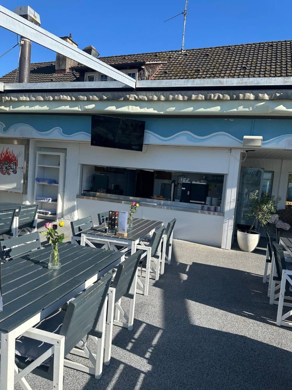 Sonnige Aussenterrasse eines Cafés mit grauen Tischen und Stühlen unter einer blauen Markise
