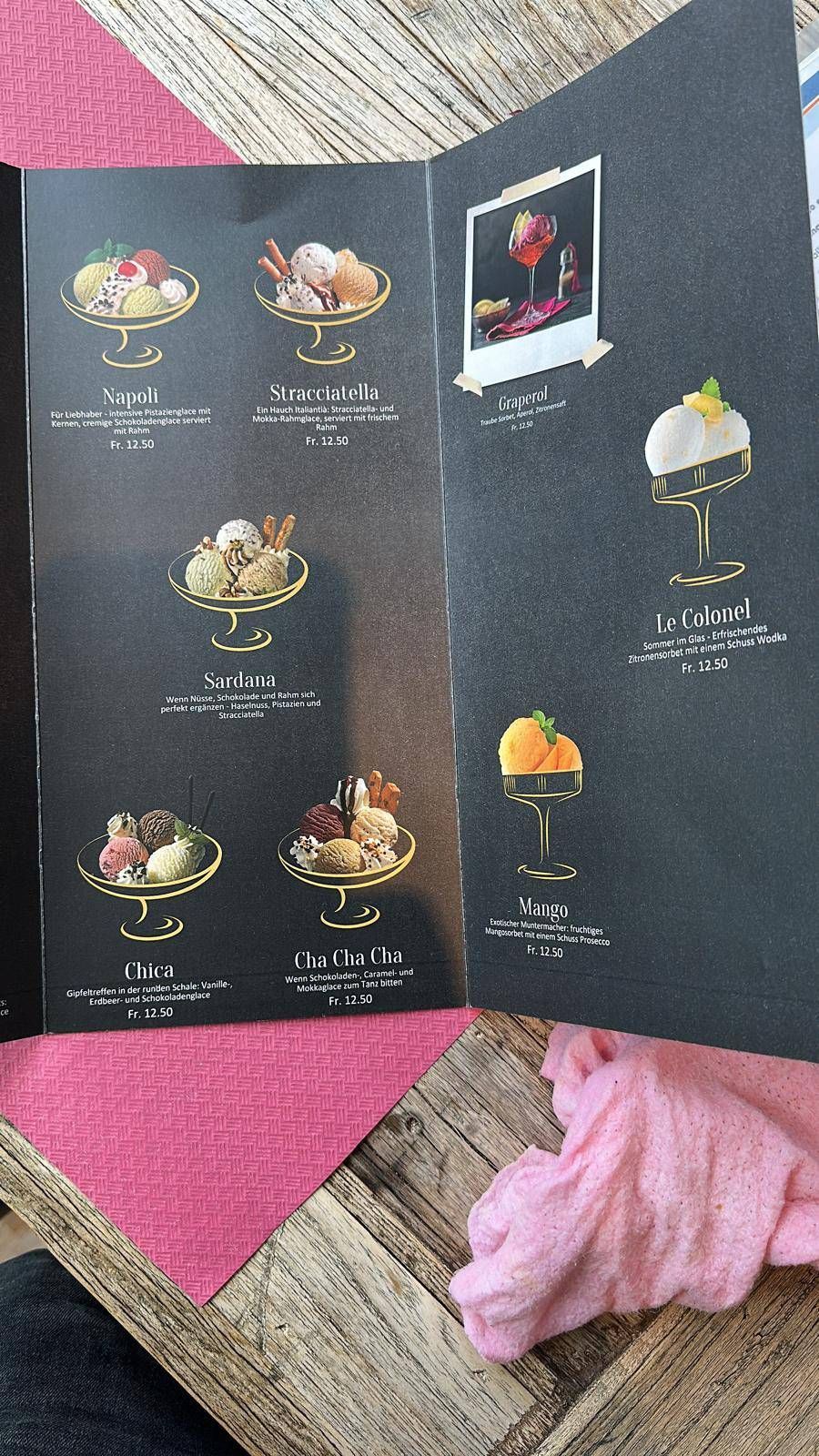 Auf einem Tisch liegt eine geöffnete Dessertkarte, auf der farbenfrohe Desserts und Getränke vor dunklem Hintergrund zu sehen sind.