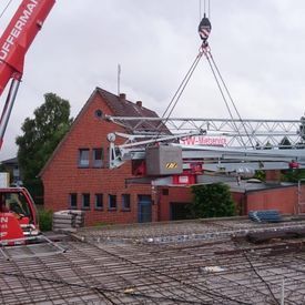 Ein großer roter Kran hebt eine Metallkonstruktion vor einem Backsteingebäude an.