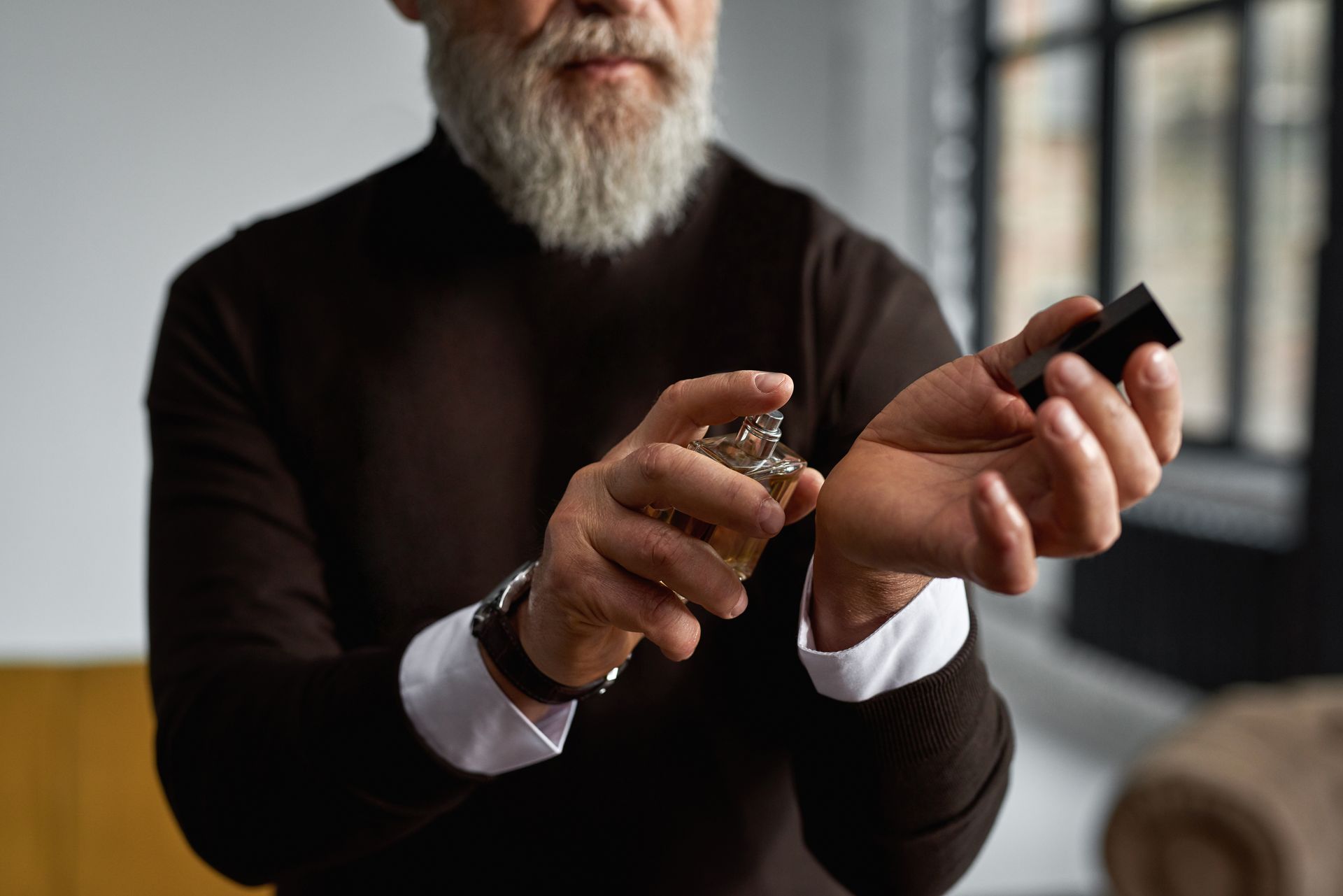 Un homme en pull sombre se parfume le poignet. Il porte une barbe blanche et une montre.
