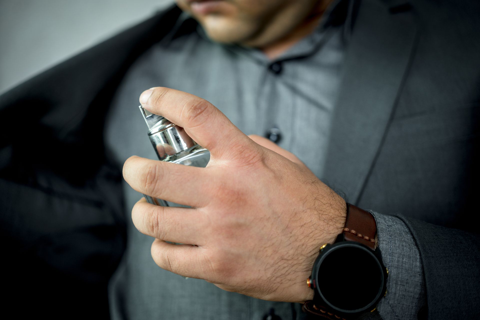 Un homme en costume gris vaporise de l'eau de Cologne sur sa poitrine, portant une montre en cuir marron.