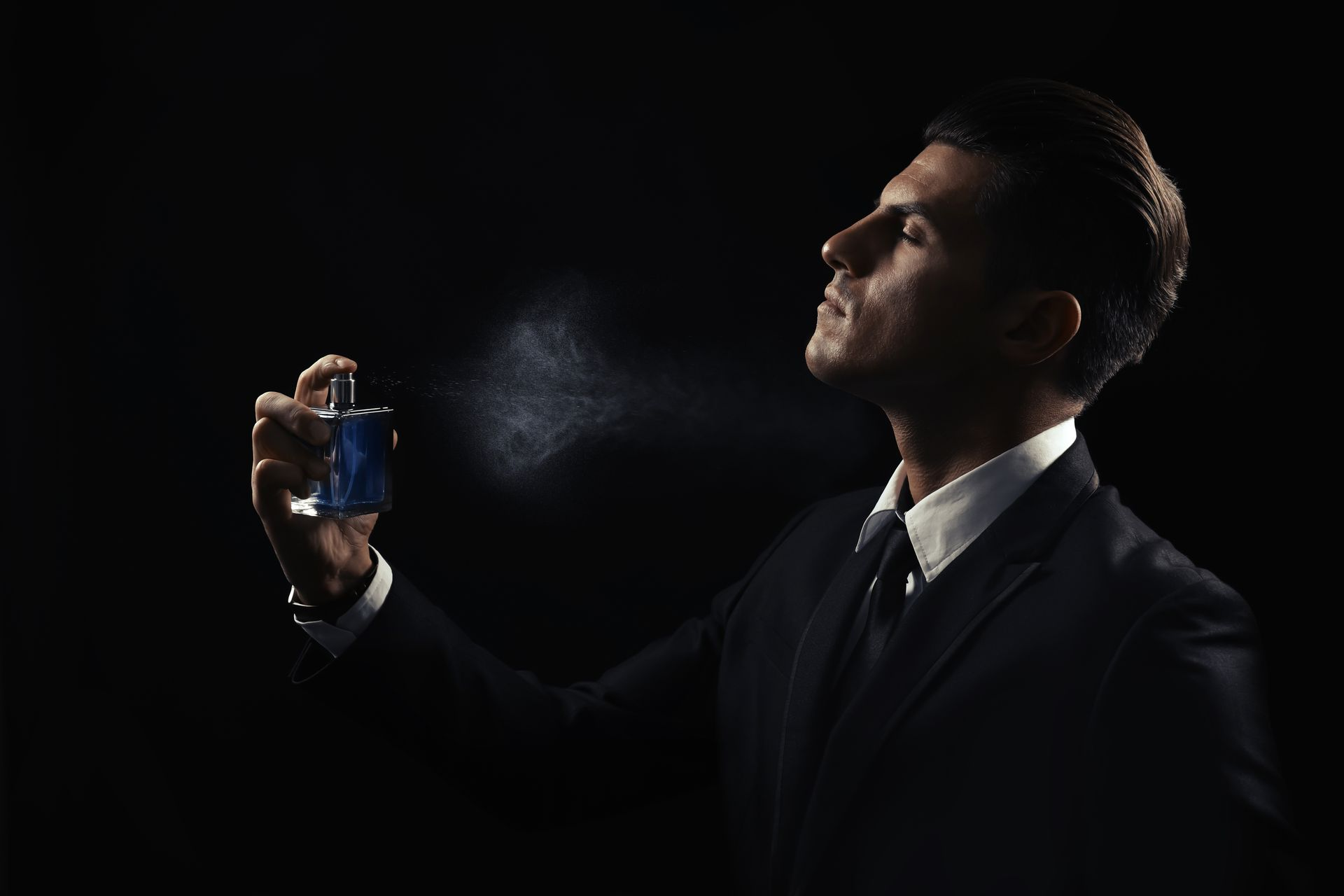 Un homme en costume vaporise du parfum, fond sombre.