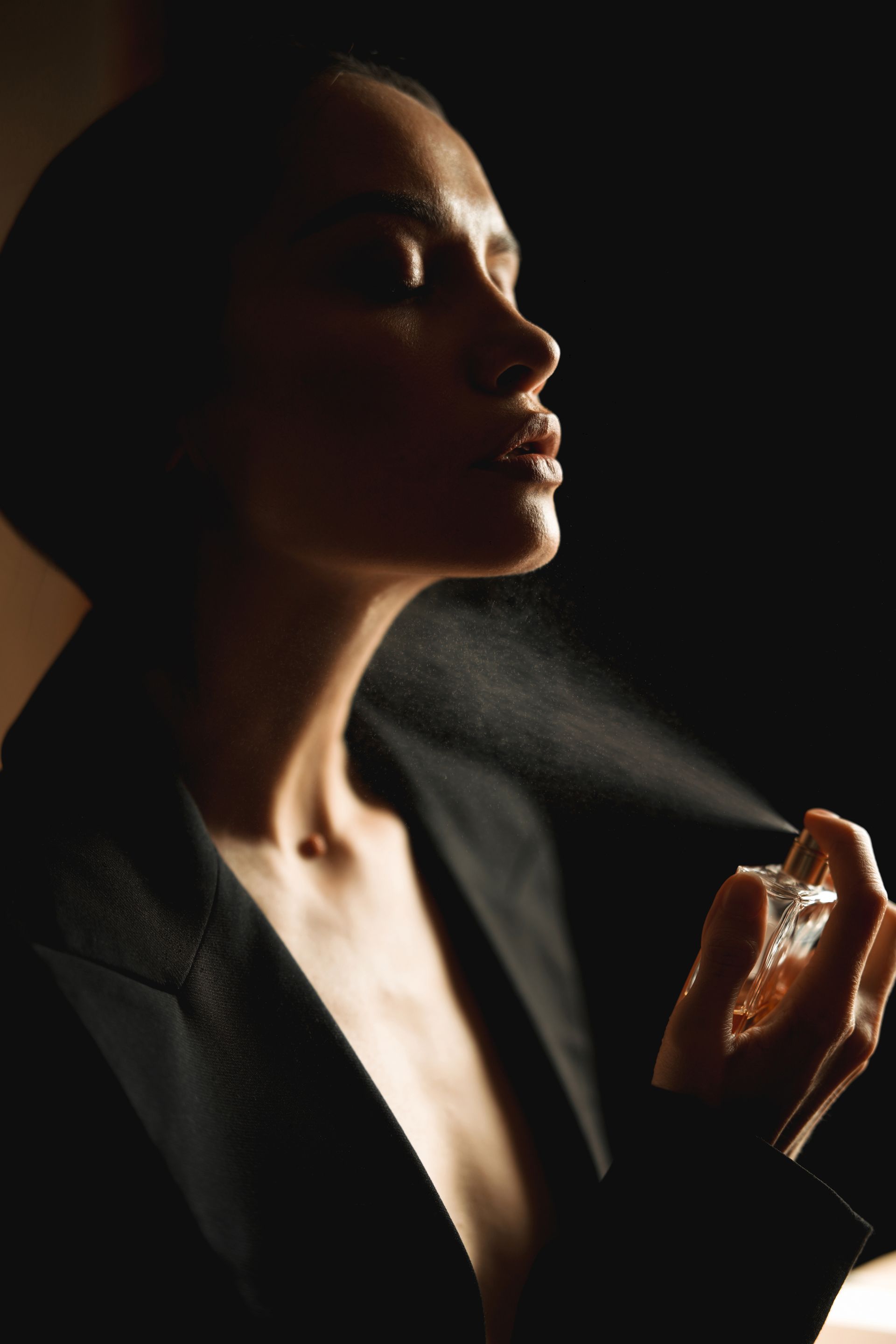 Femme en blazer se parfumant le cou. Lumière tamisée, fond sombre.