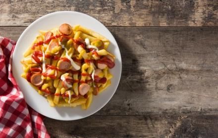 Plato de patatas fritas cubierto con salchichas en rodajas, ketchup, mostaza y mayonesa sobre una mesa de madera.