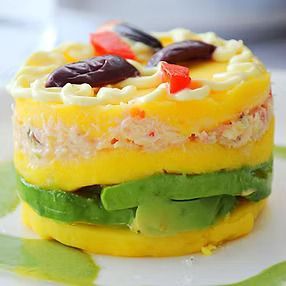 Causa peruana en capas con aguacate, cangrejo y papa amarilla. Cubierta con aceitunas y pimientos rojos.