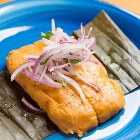 Tamal de naranja cubierto con cebolla roja y cilantro en un plato azul, sentado sobre una hoja de plátano verde.