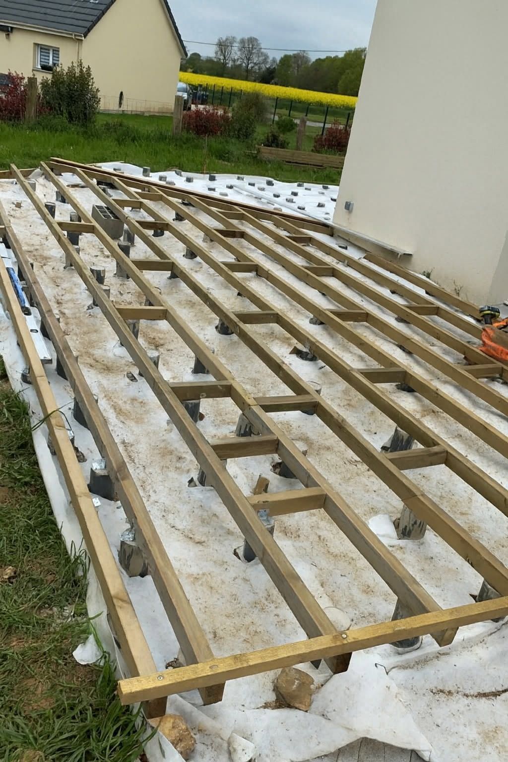 La structure d'une terrasse en bois est en construction à côté d'une maison, avec un couvre-sol blanc et un jardin en arrière-plan.