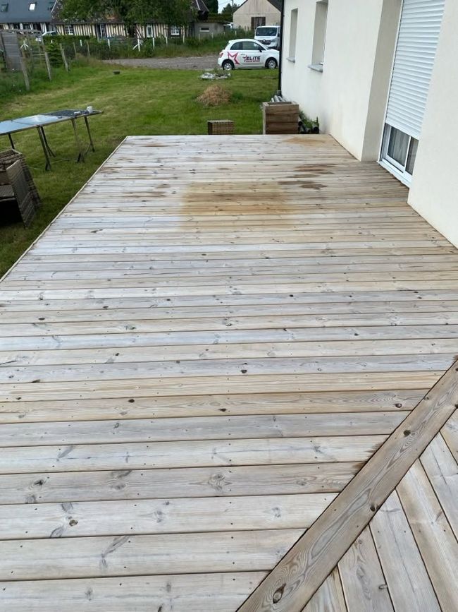 Terrasse en bois patinée donnant sur une pelouse, avec une maison en arrière-plan.