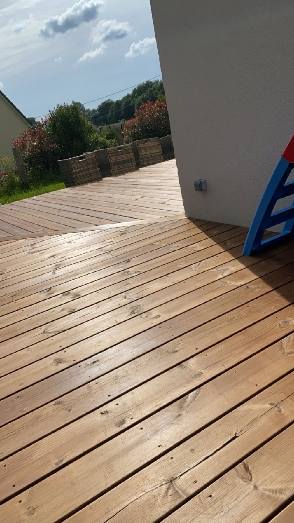 Terrasse en bois adossée au mur d'une maison, avec une échelle bleue appuyée contre celui-ci et des arbres au-delà de la clôture.