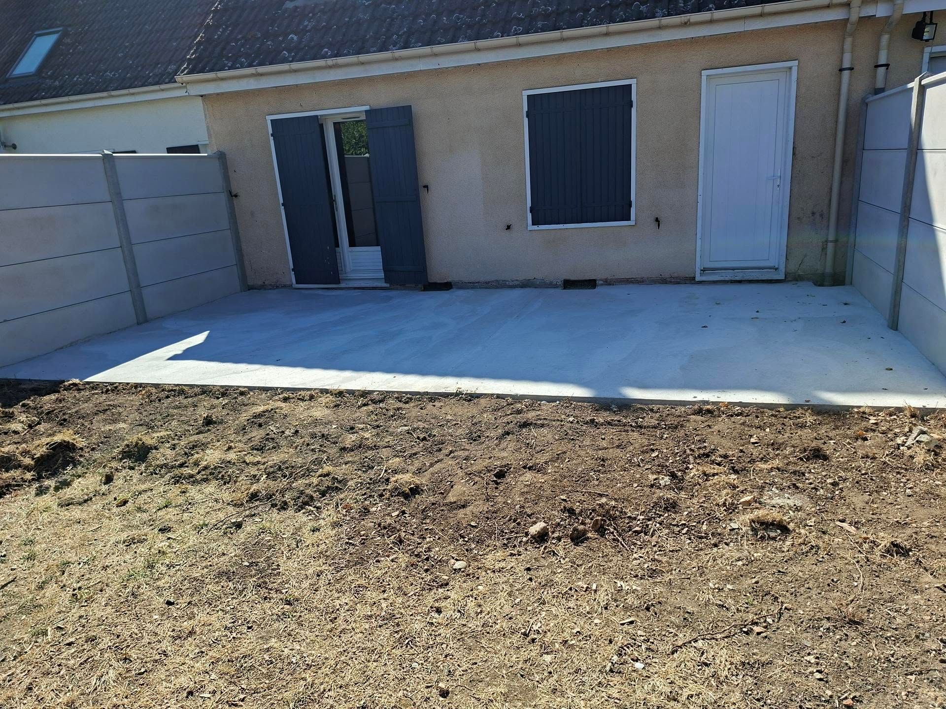 Terrasse arrière avec une dalle de béton blanc à côté d'une maison beige et d'un jardin en terre battue.