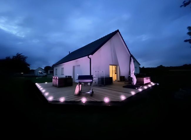 Petite maison blanche au crépuscule, avec des spots lumineux éclairant le chemin et une porte illuminée sous un ciel bleu foncé.