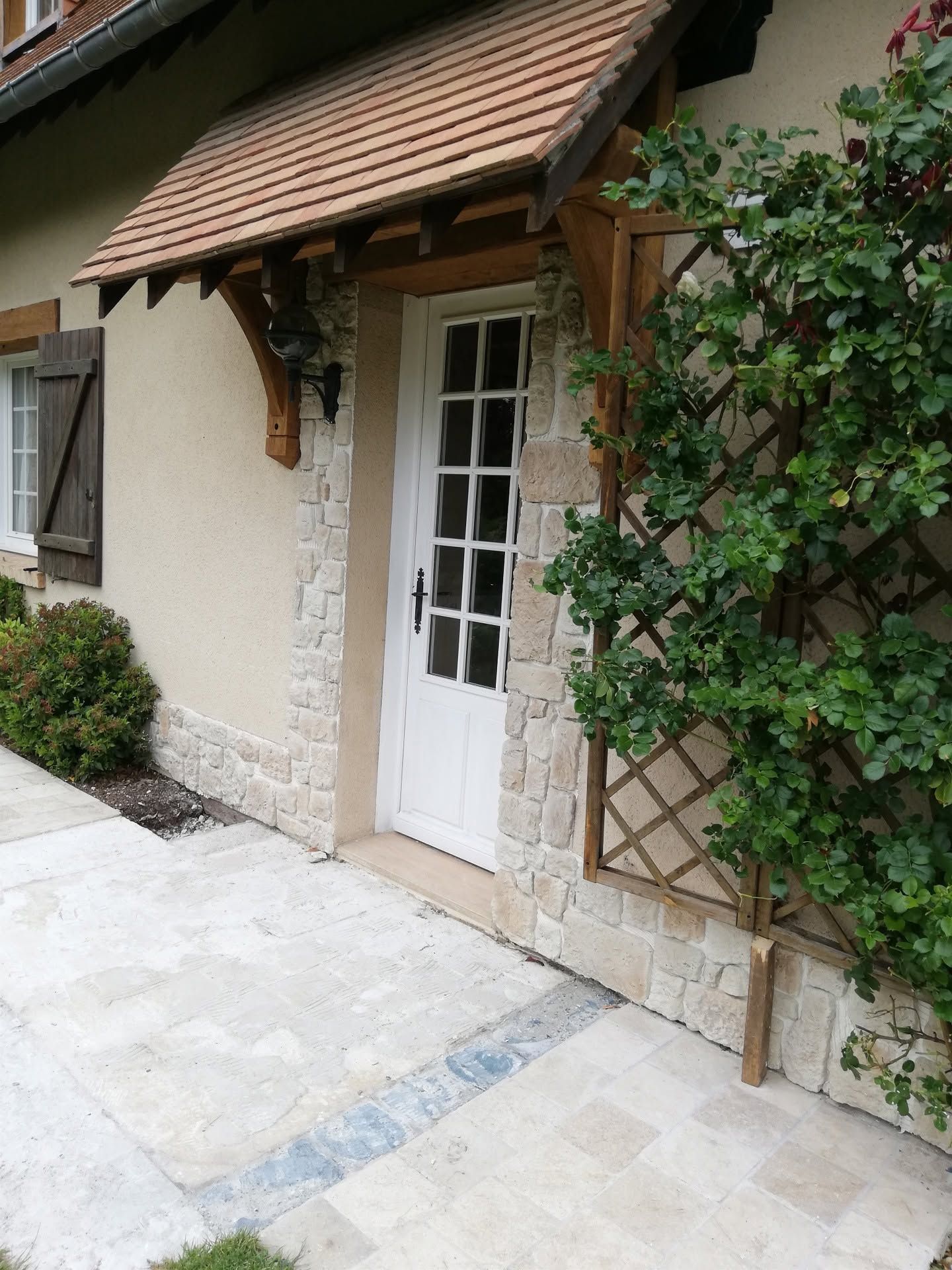 Petite entrée de maison blanche avec une porte blanche, un auvent carrelé et de la verdure luxuriante au bord d'un chemin de pierre.