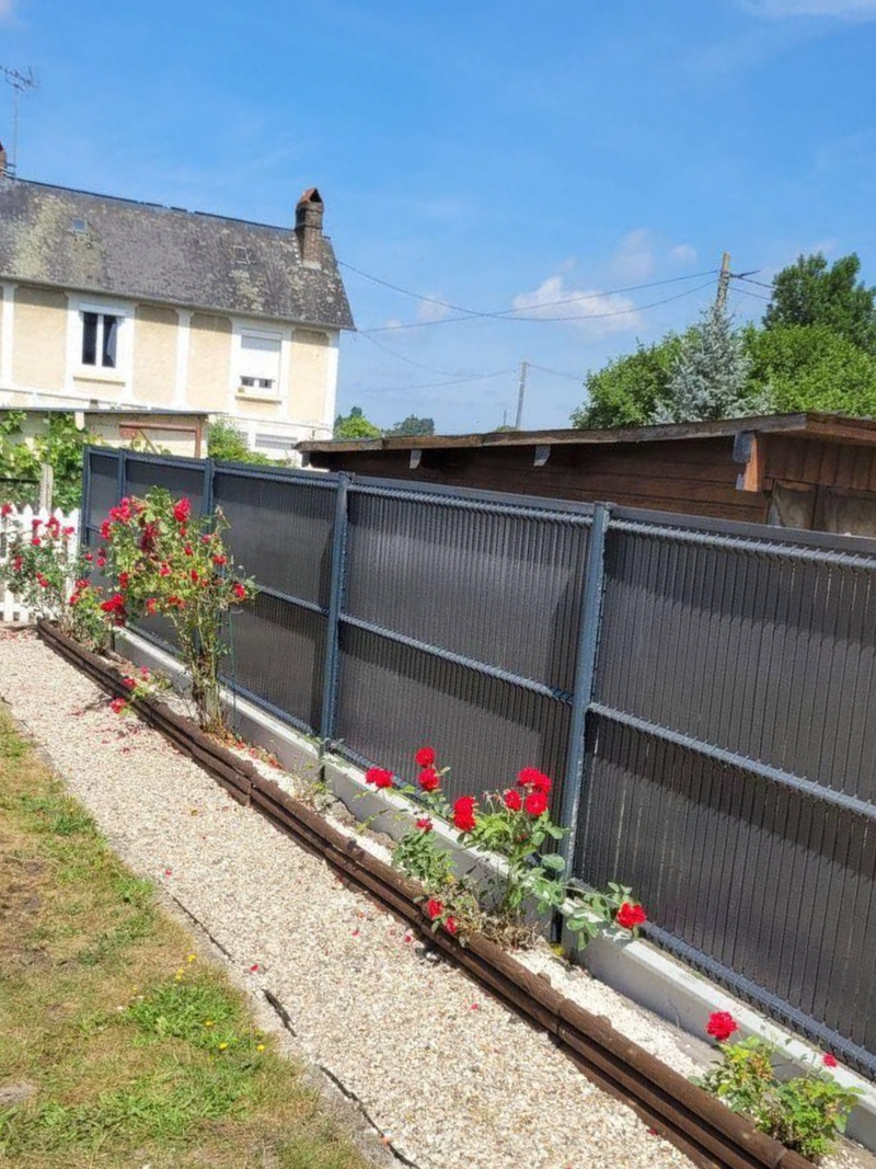 Clôture de jardin avec des fleurs rouges et une bordure de gravier à côté d'une maison par une journée ensoleillée.