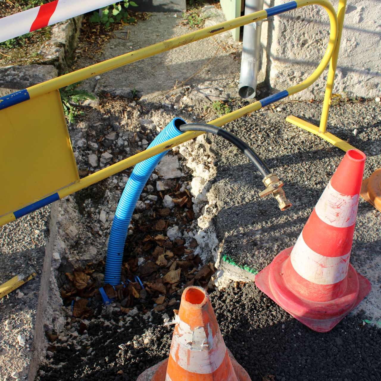Tranchée de chantier avec tuyau bleu, barrière jaune et cônes de signalisation orange sur gravier.