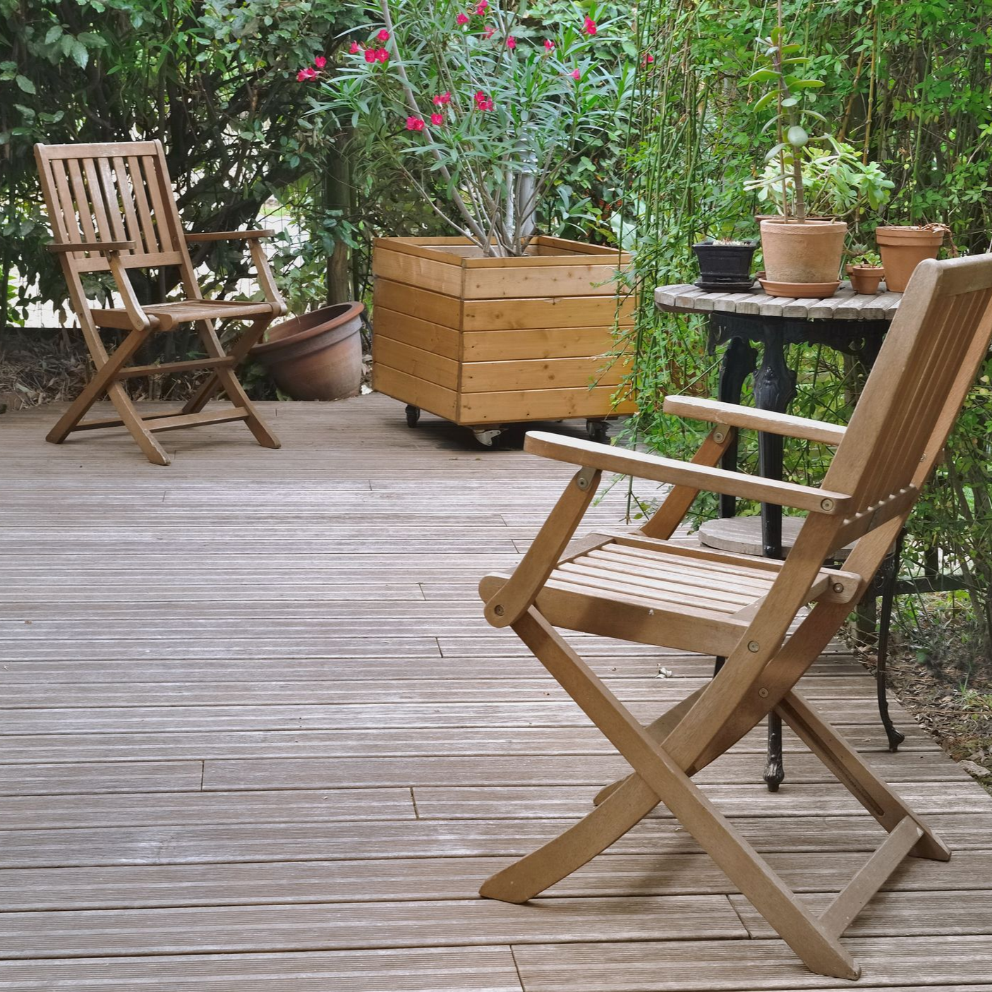 Terrasse en bois avec chaises pliantes, plantes en pot et table de jardin dans un cadre extérieur paisible.