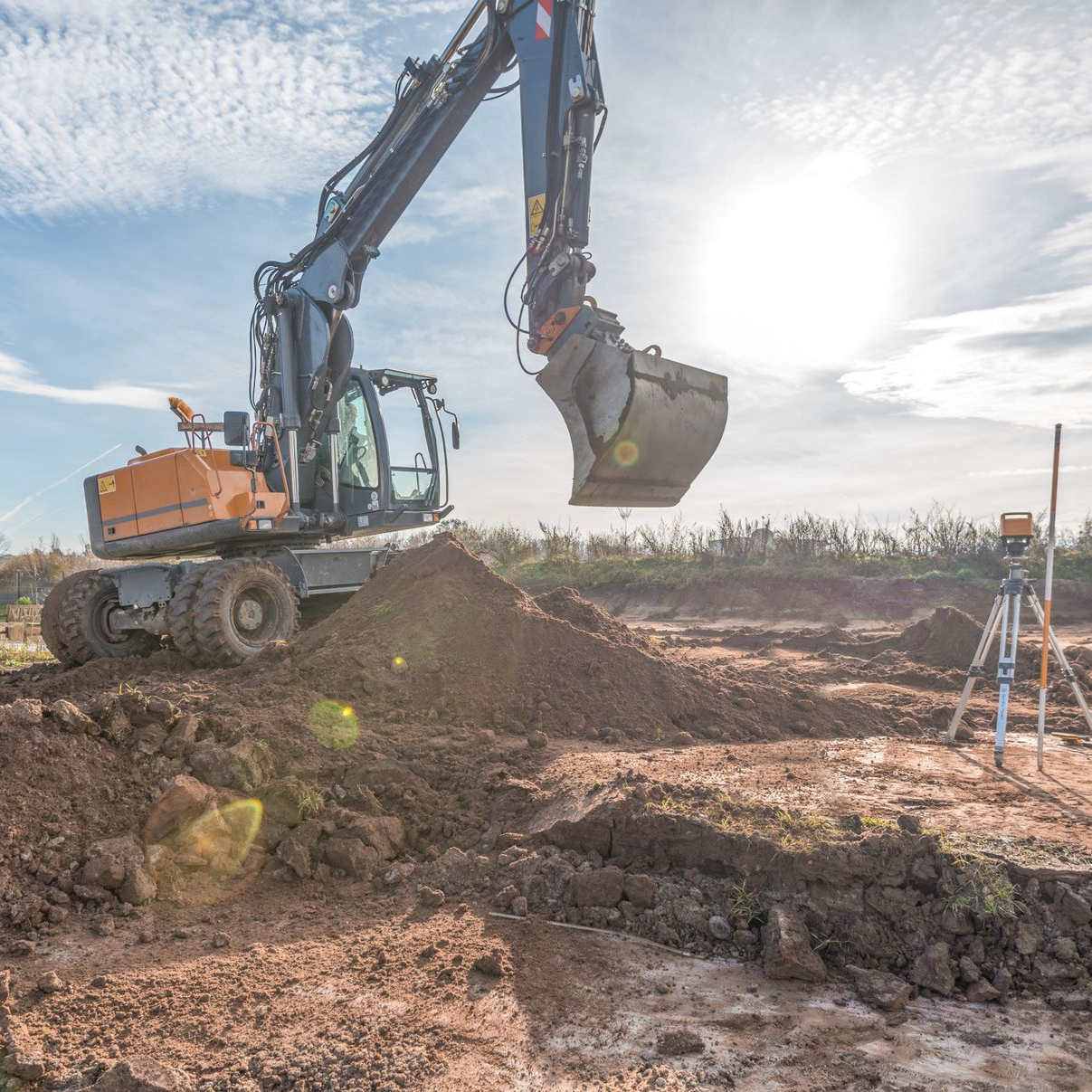 Une excavatrice sur un monticule de terre sur un chantier de construction, avec un soleil éclatant et un trépied de topographie en arrière-plan.