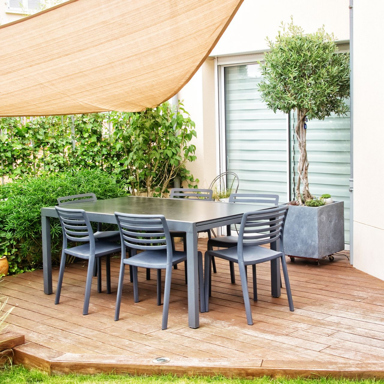 Terrasse extérieure avec une table et des chaises noires, un arbre en pot et une voile d'ombrage beige à côté d'un coin de verdure.