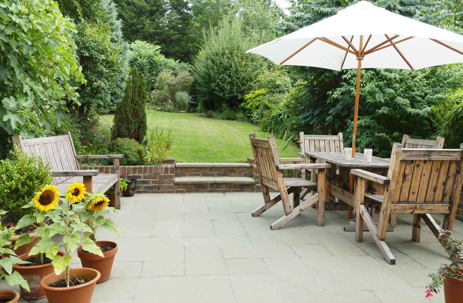 Terrasse avec table et chaises en bois sous un parasol blanc, entourée d'une végétation luxuriante et de tournesols en pot.