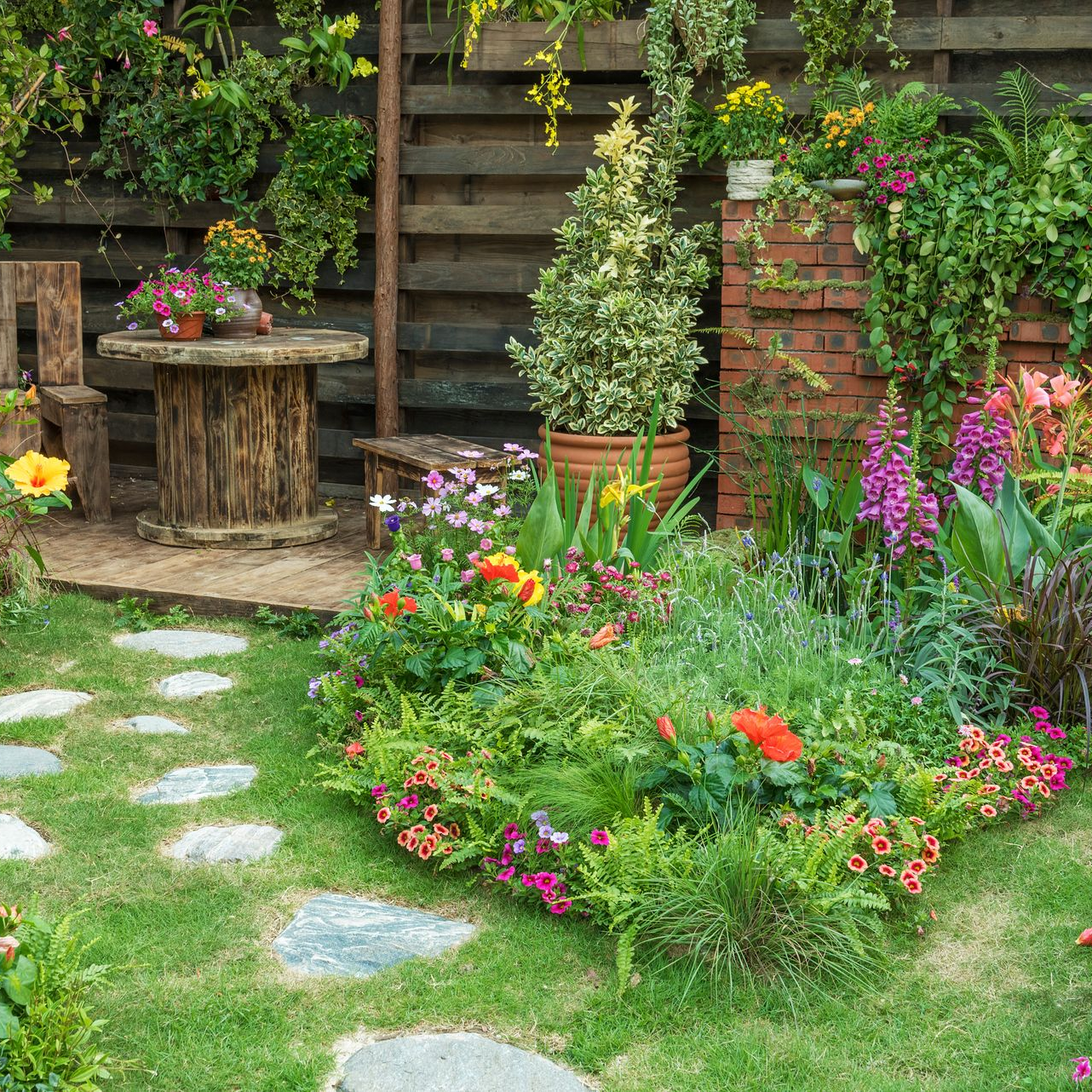Jardin fleuri et coloré avec allée de pierres, marches en bois et table rustique adossée à un mur de briques.