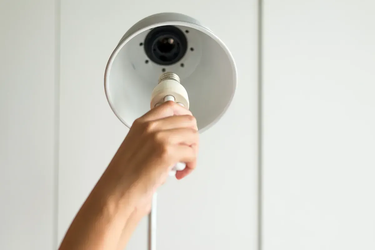 Mano de una persona enroscando una bombilla en una lámpara. La pantalla es blanca y el fondo es una pared blanca.