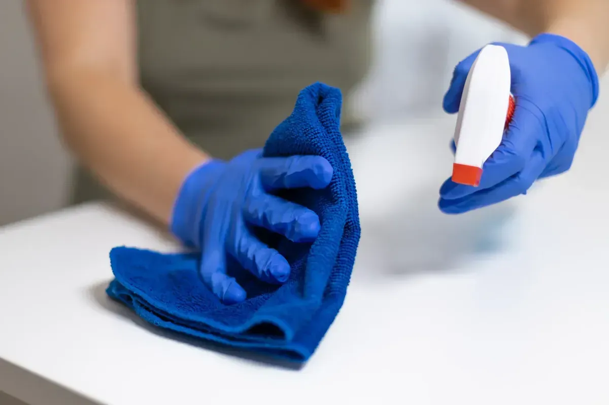 Manos con guantes azules limpiando una superficie blanca con una botella de spray y un paño azul.